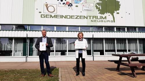 Bürgermeister Bernd Hartwig mit dem Förderbescheid und Landrätin Kerstin Weiss mit der Förderplakette vor dem Gemeindezentrum Neuburg. © LK NWM 20210326 Fördermittel Neuburg1