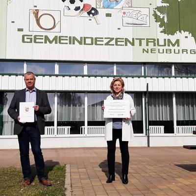Bürgermeister Bernd Hartwig mit dem Förderbescheid und Landrätin Kerstin Weiss mit der Förderplakette vor dem Gemeindezentrum Neuburg. © LK NWM 20210326 Fördermittel Neuburg1